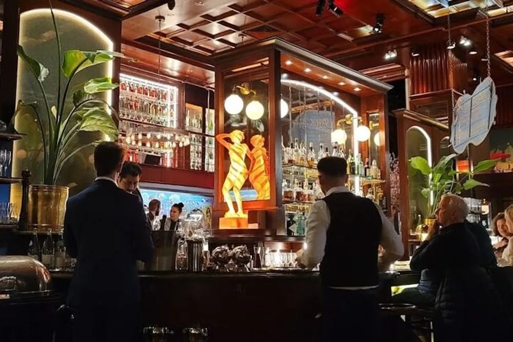 The image depicts a bustling bar scene with patrons seated bartenders at work and a warm elegant interior highlighted by a backlit artistic sculpture