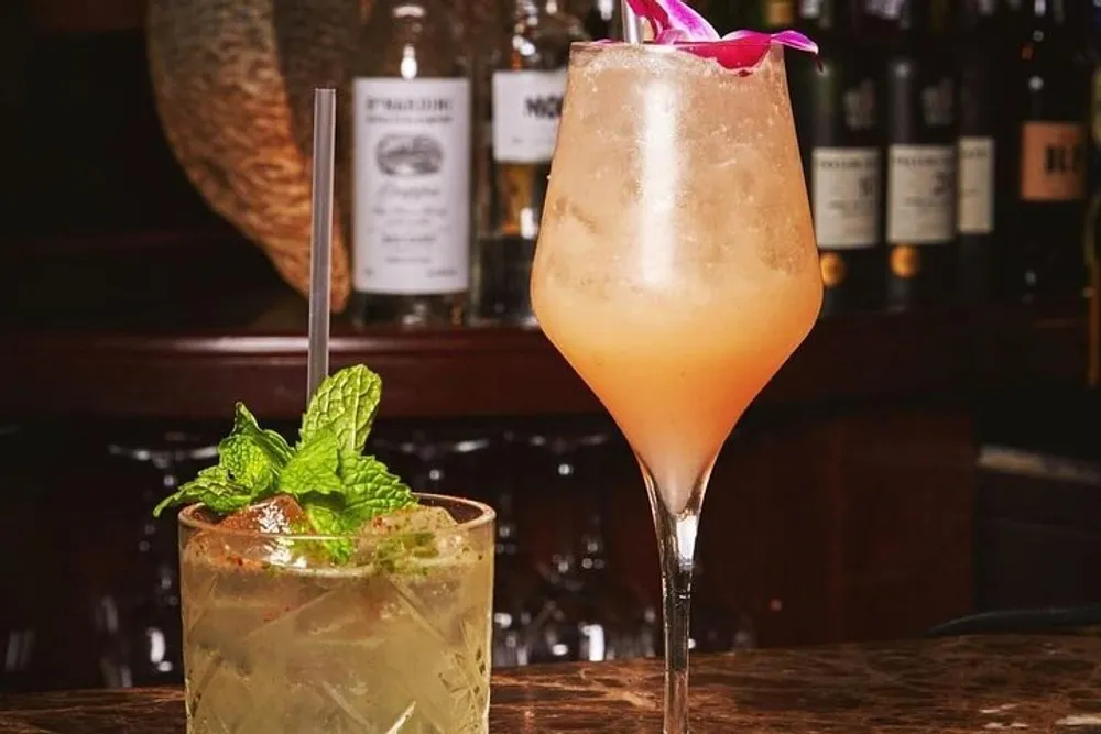 The image shows two stylishly presented cocktails on a bar counter with one garnished with mint and the other with a pink flower