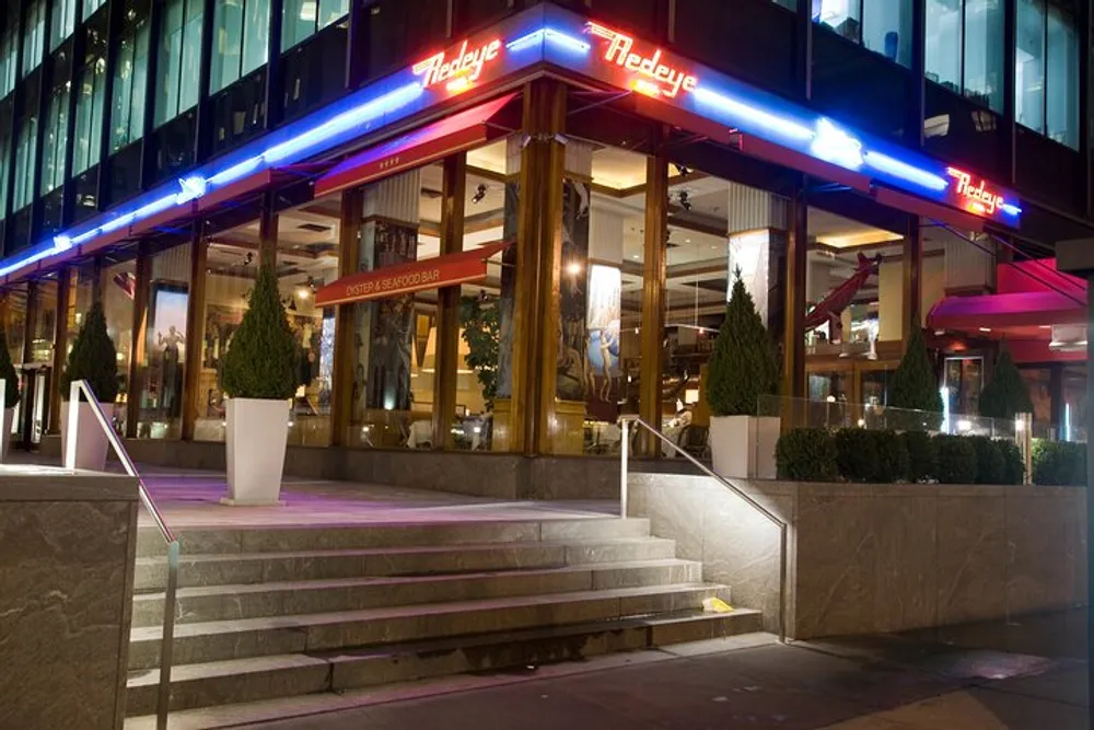 The image displays the exterior of a modern restaurant named Redeye Grill at nighttime illuminated by neon lighting above the entrance