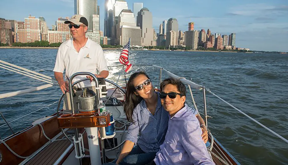 A couple is enjoying a sailboat ride on a sunny day with a city skyline in the background while a skipper steers the boat
