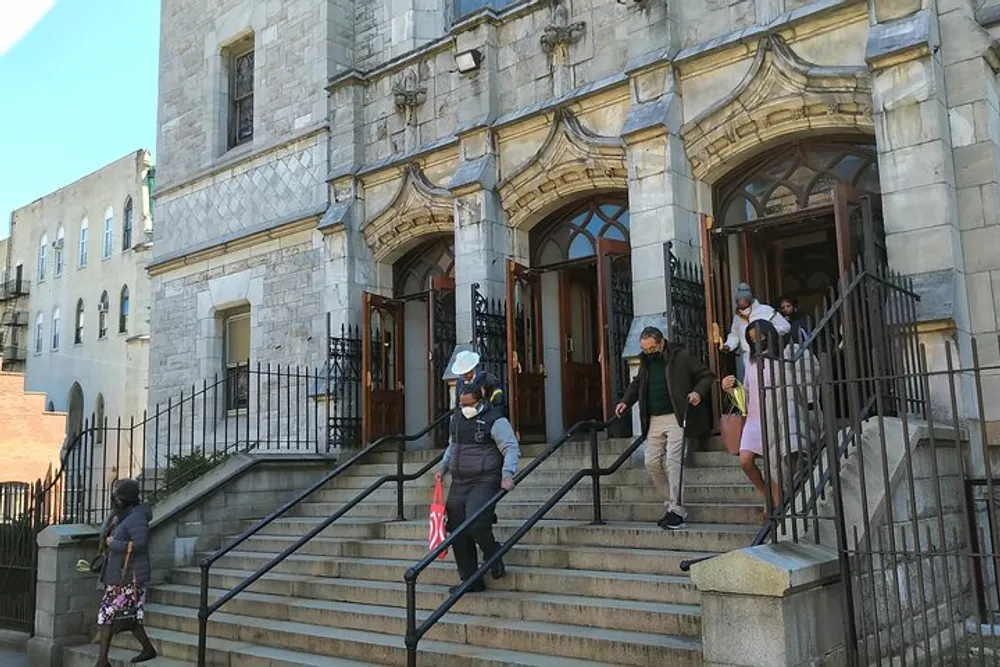 People are descending the steps of a stone-faced building with ornate doors implying that they may have just attended an event inside