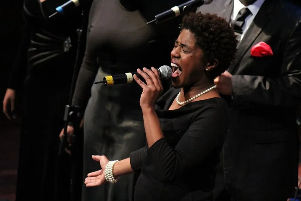A woman passionately sings into a microphone with her eyes closed as part of a choir or music group performance