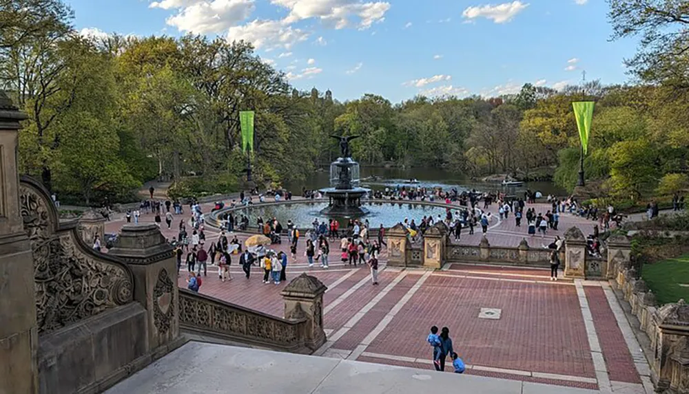 The image shows a bustling outdoor scene with people gathering around a fountain in a park with green trees and blue sky in the background