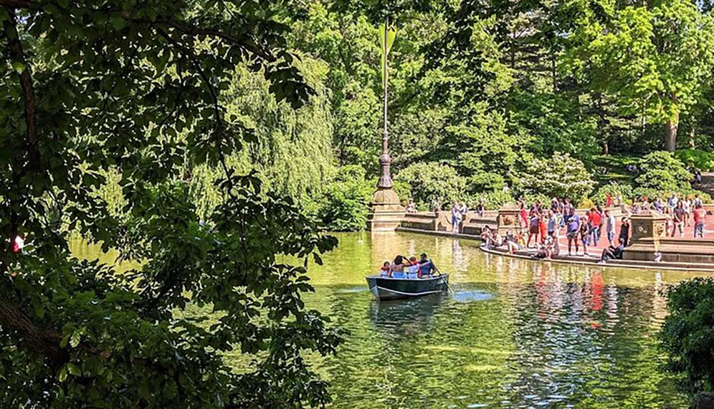 The image shows a serene park setting where people are enjoying a sunny day by rowing a boat on a calm pond and walking along its banks under lush green trees