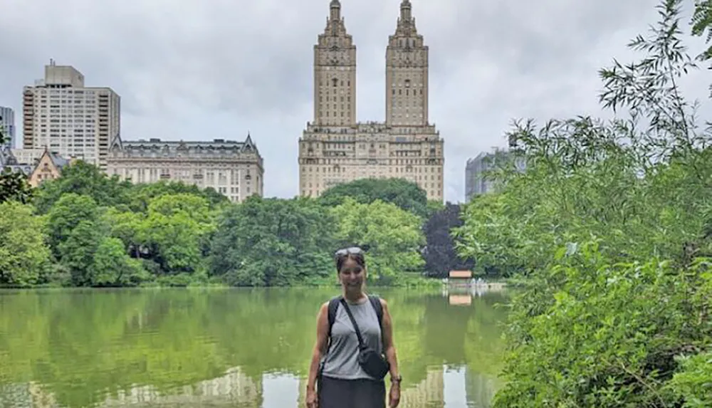 A person is standing in front of a serene pond with towering buildings in the background likely in a city park