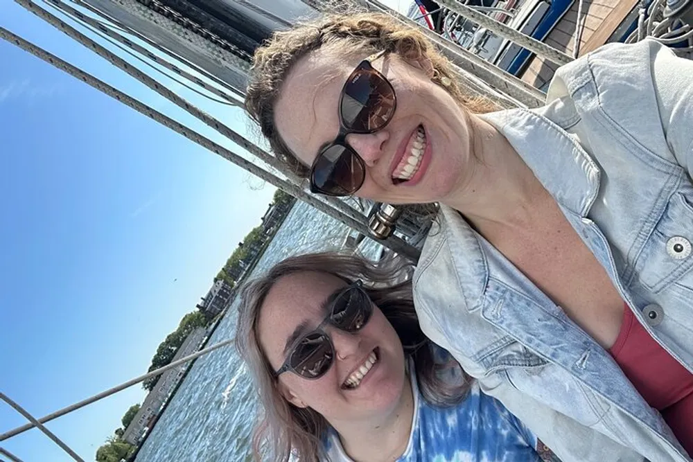 Two smiling women wearing sunglasses are taking a selfie on a bright sunny day with rigging lines from a boats mast visible in the background suggesting they are on or near a sailboat
