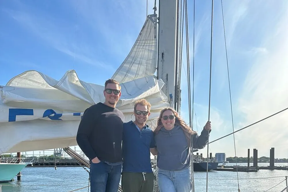 Three people are smiling together in front of a partially folded sail on a sunny day at the marina