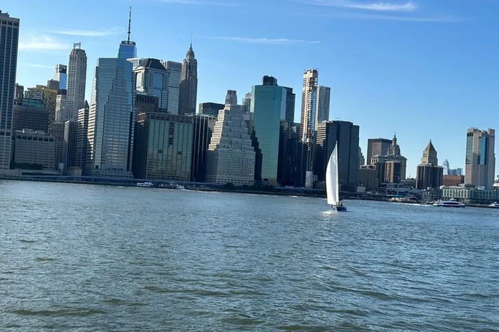 The image shows a sailboat on the water with the skyline of Lower Manhattan New York City in the background on a sunny day