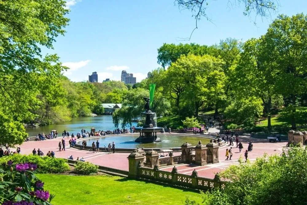 The image shows a vibrant and bustling central area in a park with a fountain surrounded by lots of greenery and people enjoying a sunny day