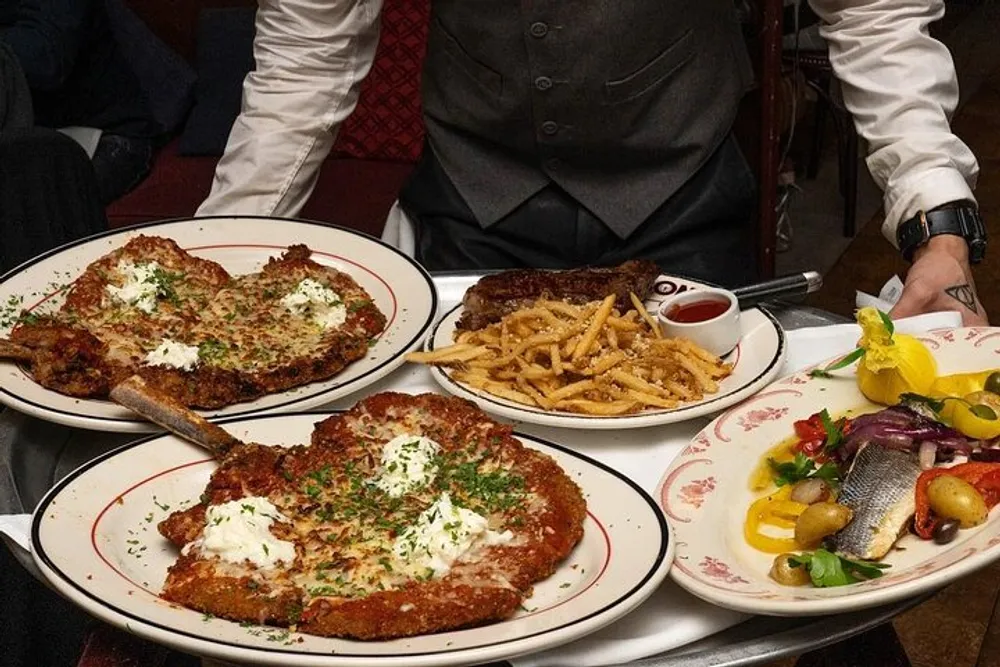 A waiter is serving large portions of different dishes such as breaded meat cutlets and fries on plates at a dining table