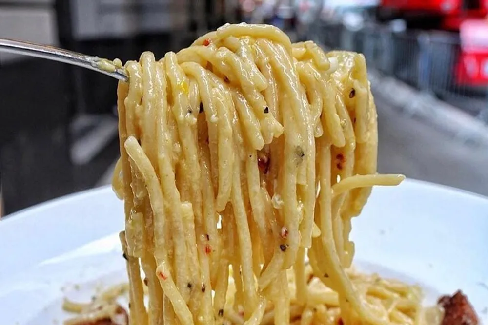 A close-up of a fork lifting a creamy pepper-flecked spaghetti likely a type of pasta like cacio e pepe with a blurry street scene in the background