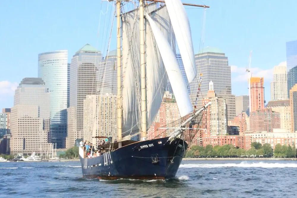 A tall ship with white sails is cruising on the water with a city skyline in the background