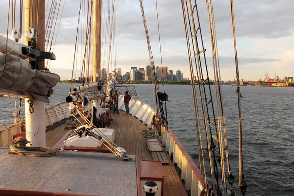 A view from the deck of a tall ship with its rigging visible sailing towards a city skyline during sunset