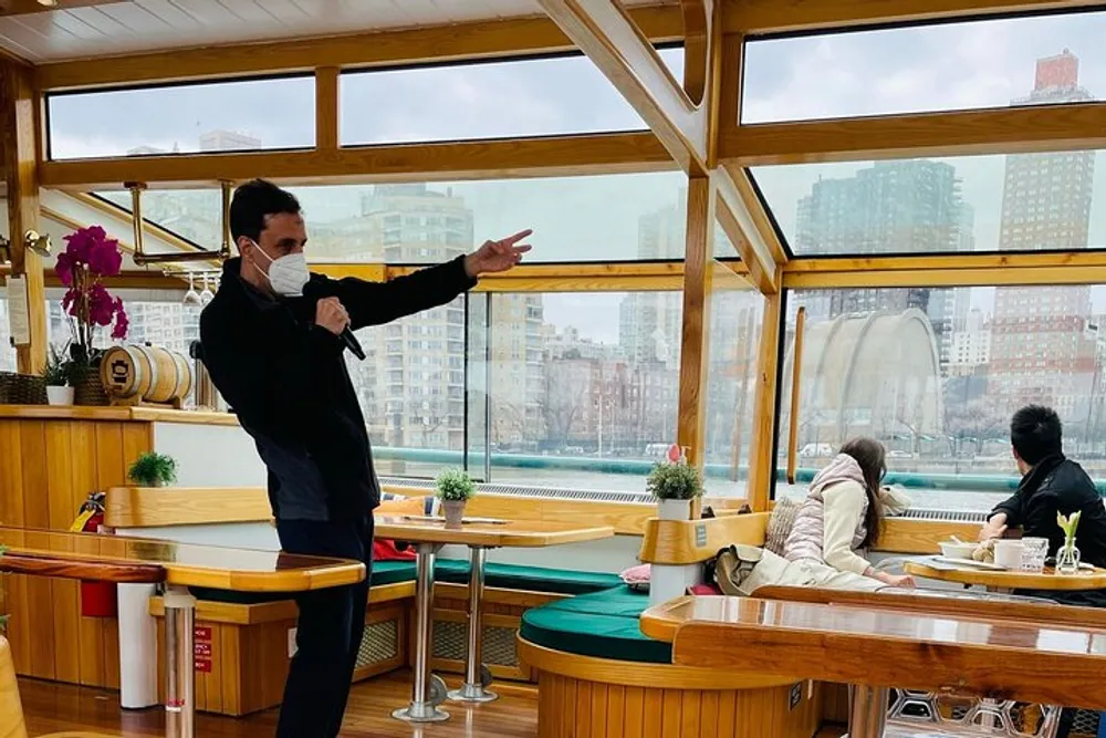 A masked individual is pointing towards something outside the window while addressing others seated inside a boat with wooden interiors and modern cityscape views