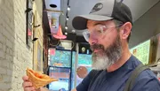 A man with a beard wearing a baseball cap and safety glasses looks at a slice of pizza he is holding.