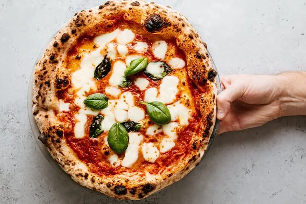 A hand is holding a freshly baked pizza topped with melted mozzarella basil leaves and a rich tomato sauce with some charred spots on the crust indicating it may have been cooked in a wood-fired oven