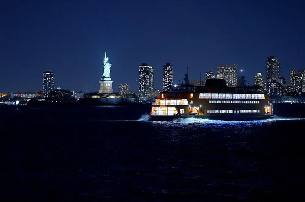 A brightly lit ferry passes by the Statue of Liberty against the backdrop of a twinkling city skyline at night