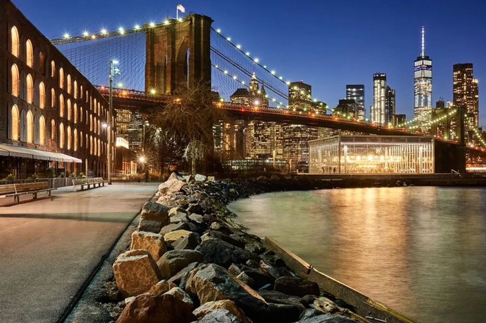 The image captures a serene evening view of the Brooklyn Bridge with the illuminated New York City skyline in the background