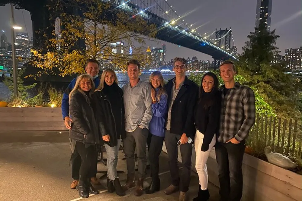 A group of smiling people pose for a photo at night with a lit-up bridge and city skyline in the background