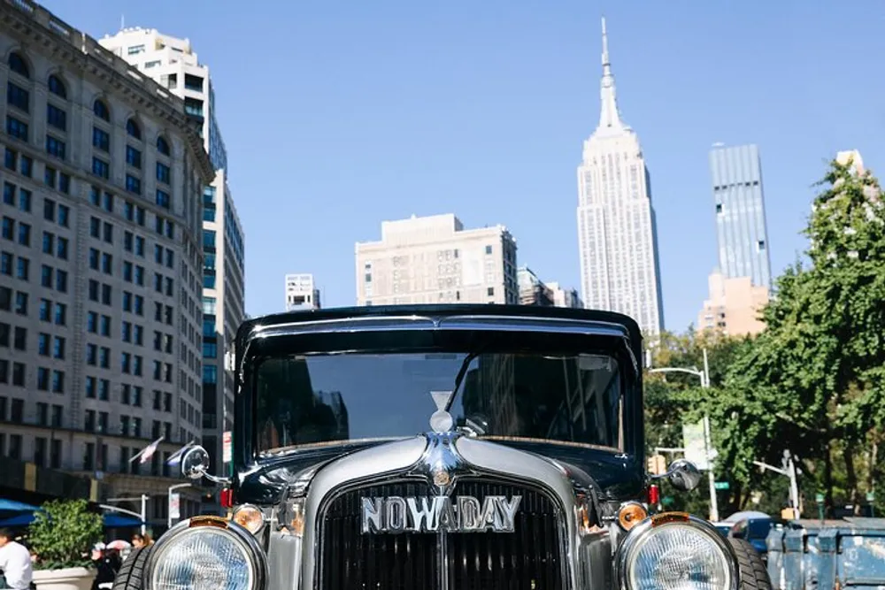 A vintage car with the license plate NOWADAY is parked with the Empire State Building visible in the background