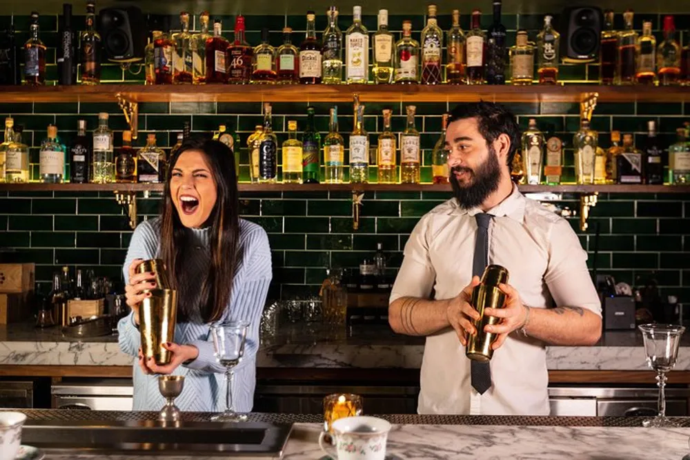 Two bartenders are sharing a lively moment behind a bar stocked with various bottles of alcohol with one of them laughing heartily while shaking a cocktail shaker