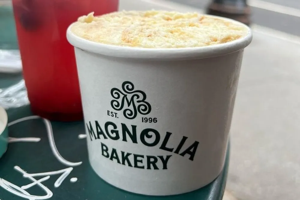 A paper container with the Magnolia Bakery logo contains a dessert juxtaposed with a red drink and a napkin in the background