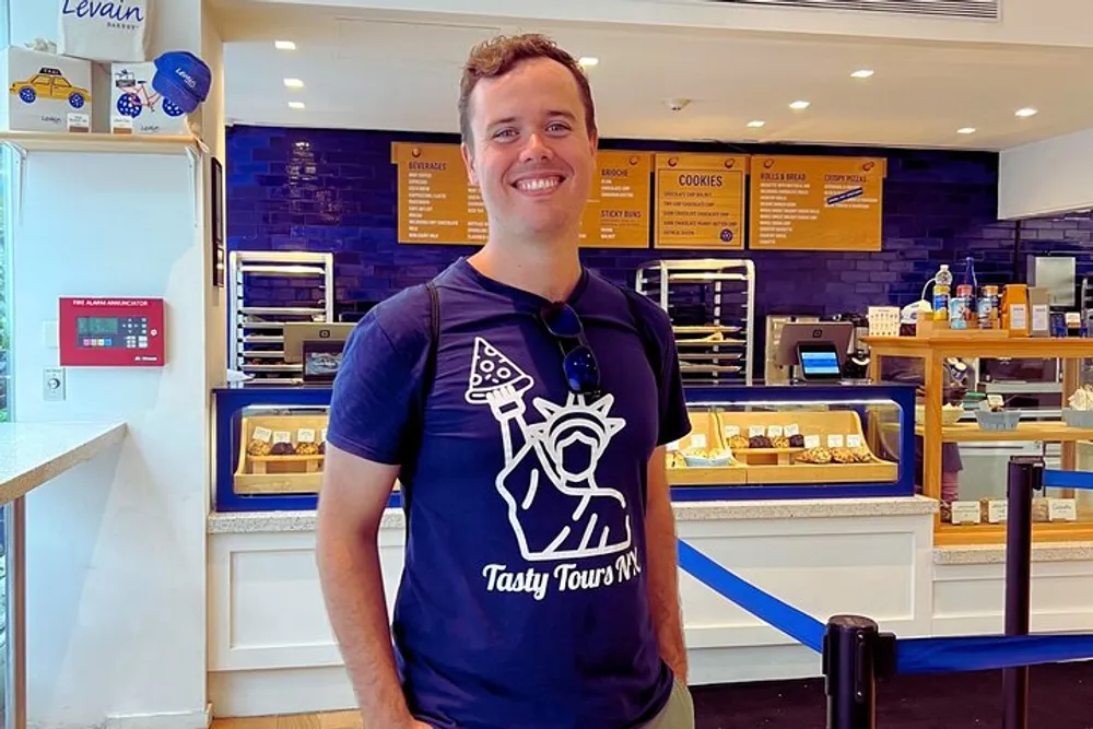 A smiling man is standing inside a bakery with a blue and white interior in front of a counter displaying various baked goods
