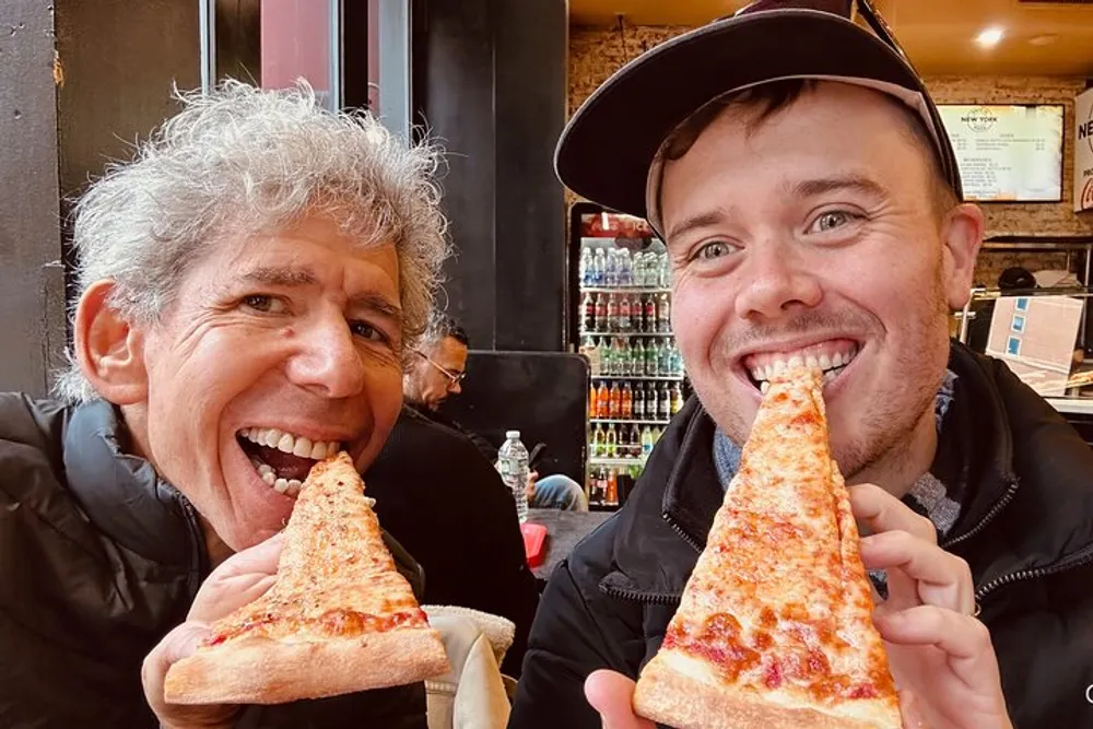 Two people are happily posing for a selfie with large slices of pizza in what appears to be a casual dining setting