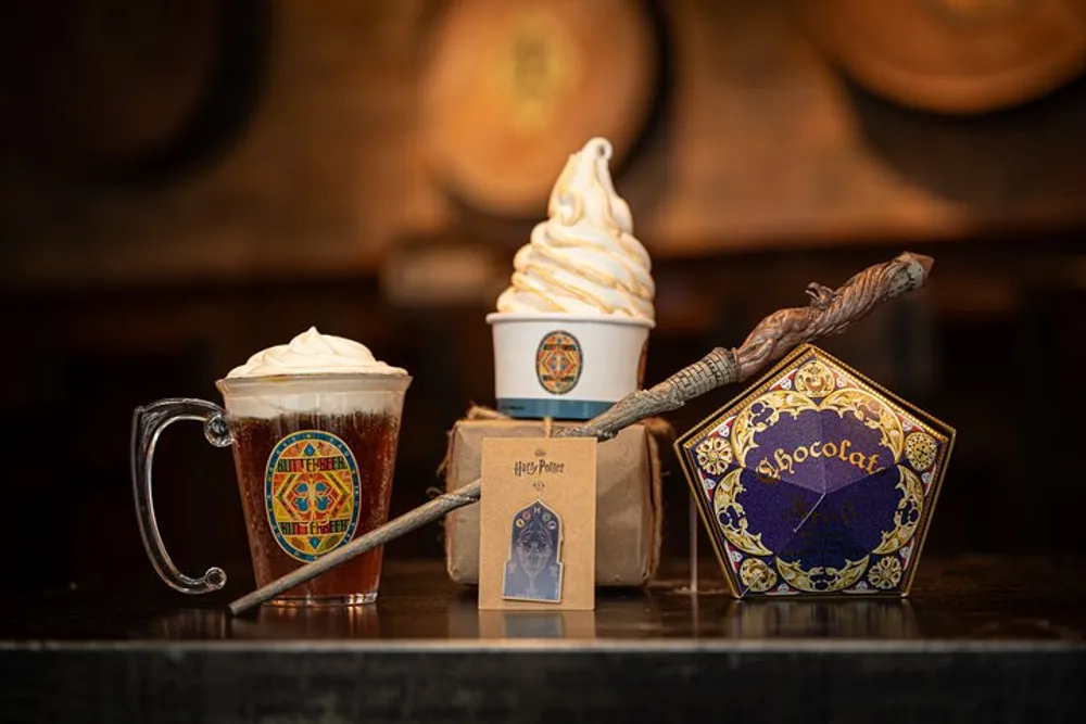 The image features an assortment of Harry Potter-themed items including a butterbeer in a mug with whipped cream on top a soft-serve ice cream cone a chocolate frog package a wand and a brown paper bag with a logo all set against the backdrop of barrels