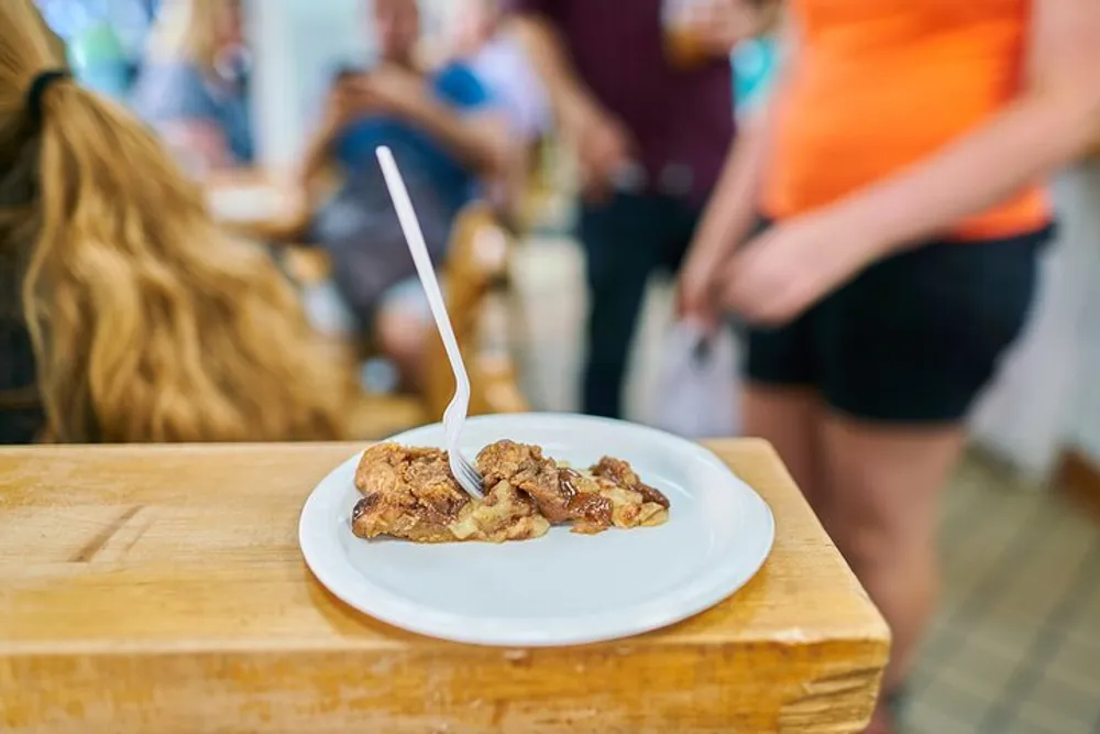 A plate of food with a plastic fork is sitting on a wooden counter in a room with people in the background