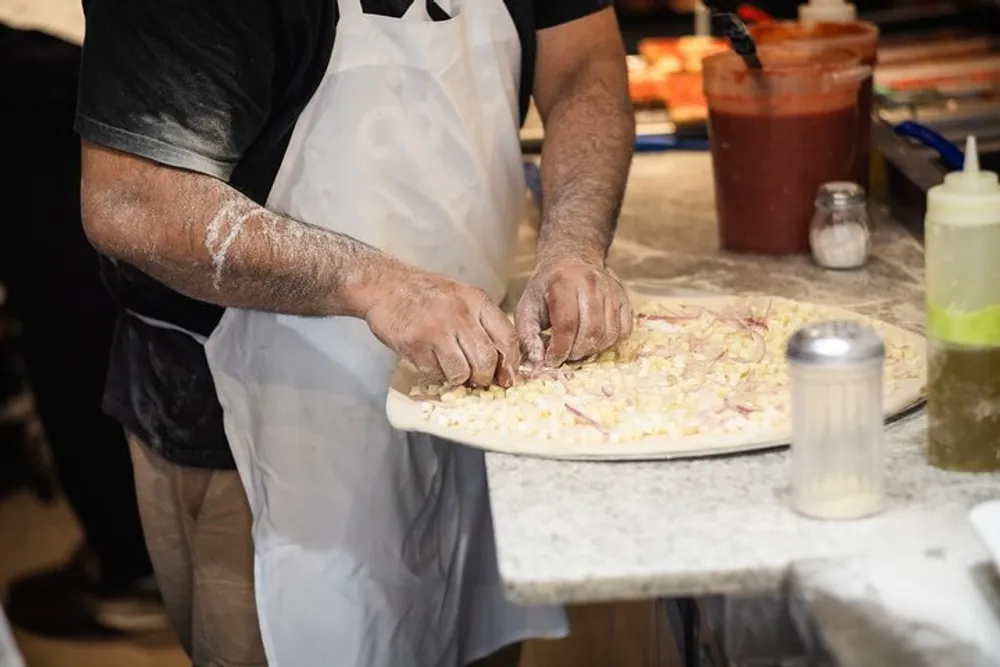 A person wearing an apron is preparing food with their hands working on what appears to be a pizza topped with onions amidst a kitchen setting with ingredients and cooking utensils around