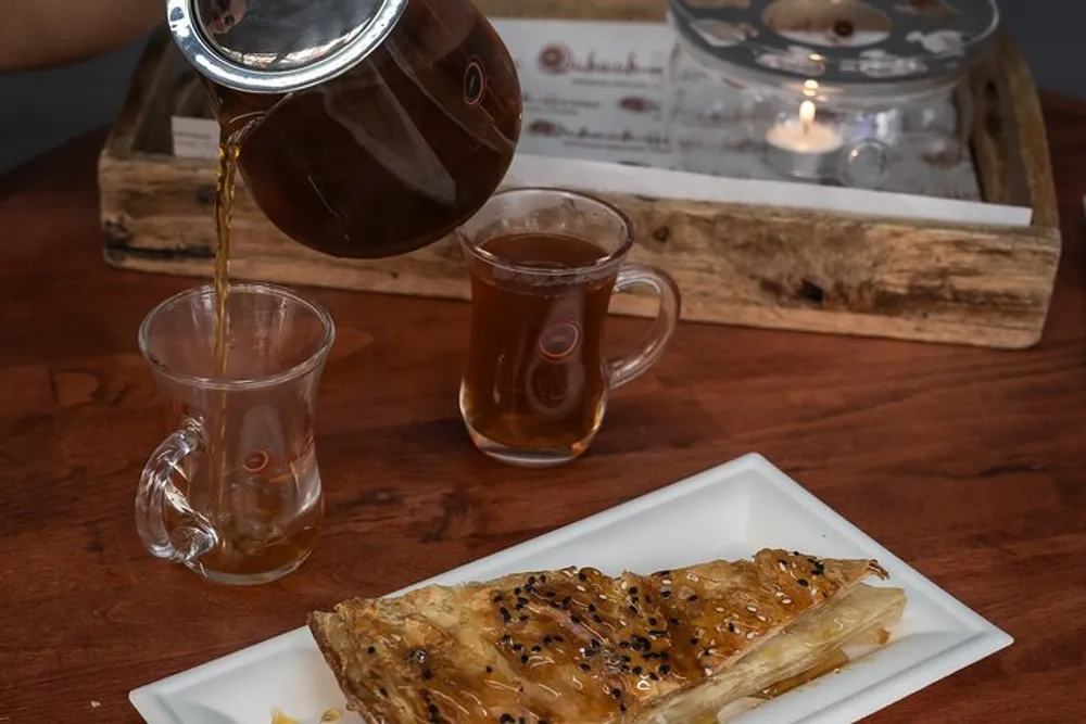 Tea is being poured from a teapot into a clear glass cup next to another filled cup and a plate with a piece of baklava on a table with a lit candle in the background
