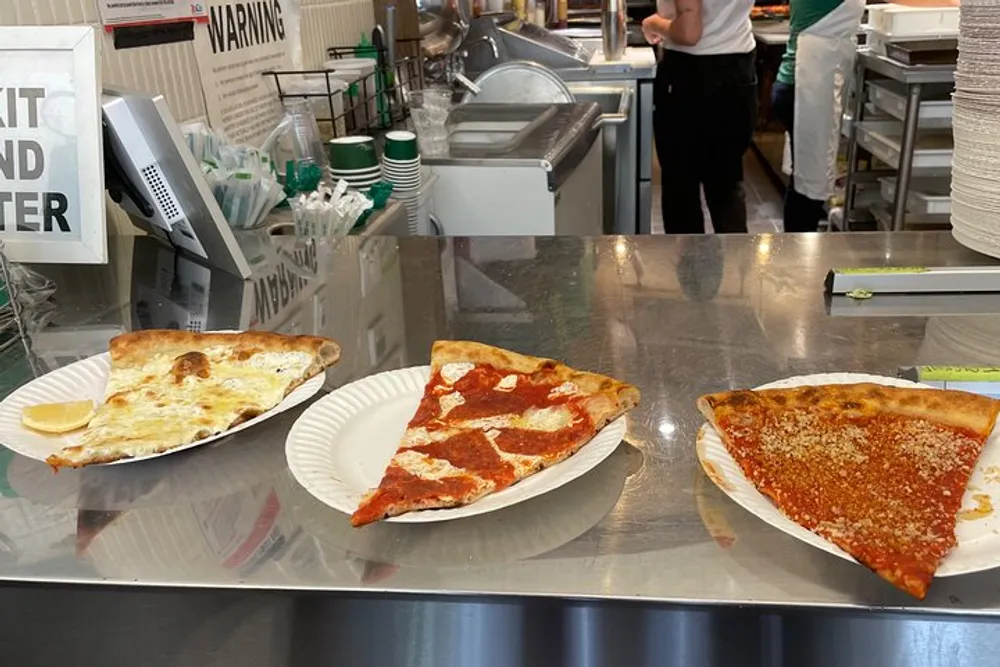 Three slices of pizza with different toppings are presented on paper plates on a counter in a pizzeria with a person visible in the background