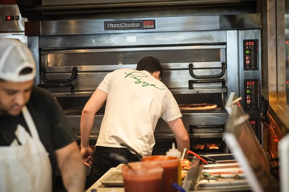 A person wearing a white t-shirt is working at a pizza oven while another person in an apron observes from the foreground