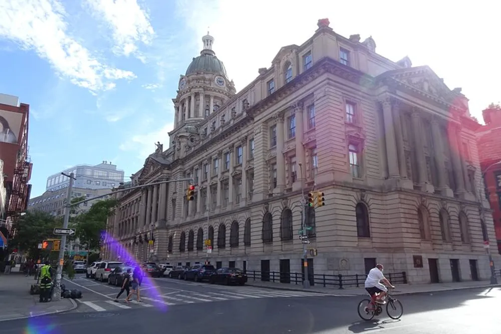 The image captures a sunlit street corner with a grand historic building traffic lights pedestrians crossing the road and cyclists under a clear blue sky