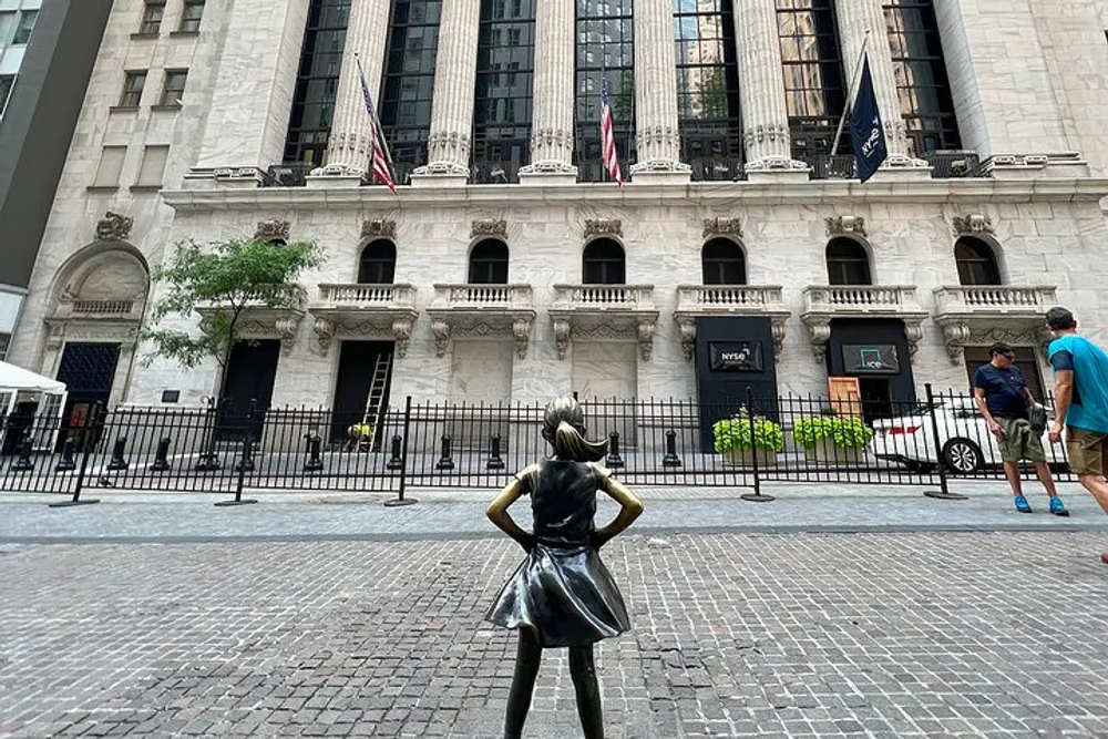 A bronze statue of a girl stands with her hands on her hips facing the New York Stock Exchange with passersby and American flags in the background