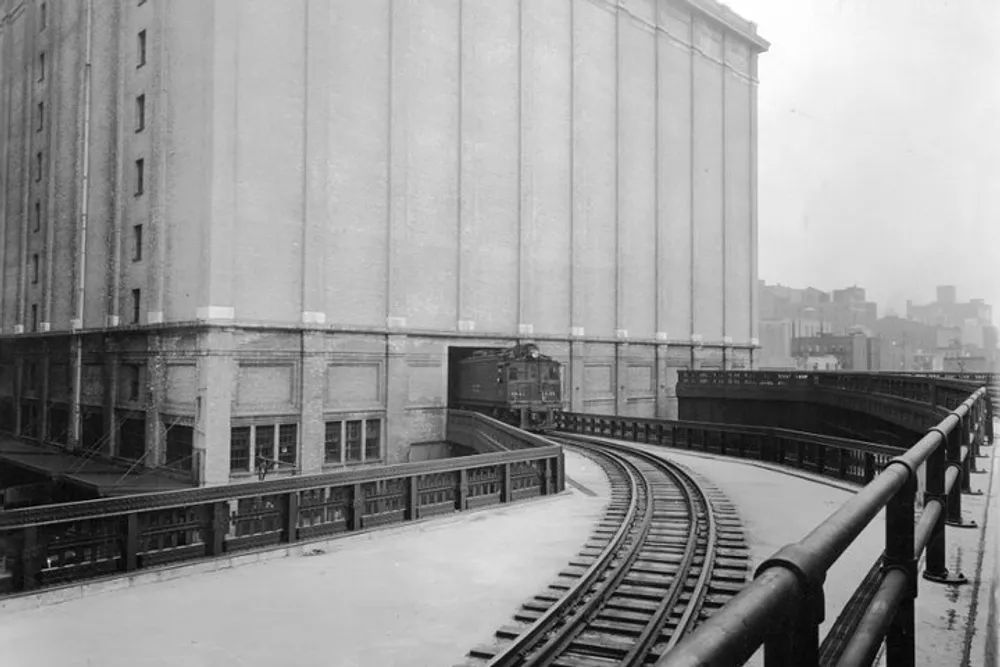 The image shows an elevated train line curving into a tunnel beneath a large industrial or storage building evoking an early to mid-20th century urban setting
