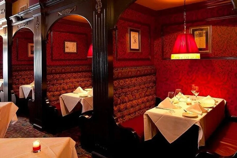 The image shows a cozy and elegant restaurant interior with red walls patterned upholstery white tablecloths and warm lighting from red-shaded lamps