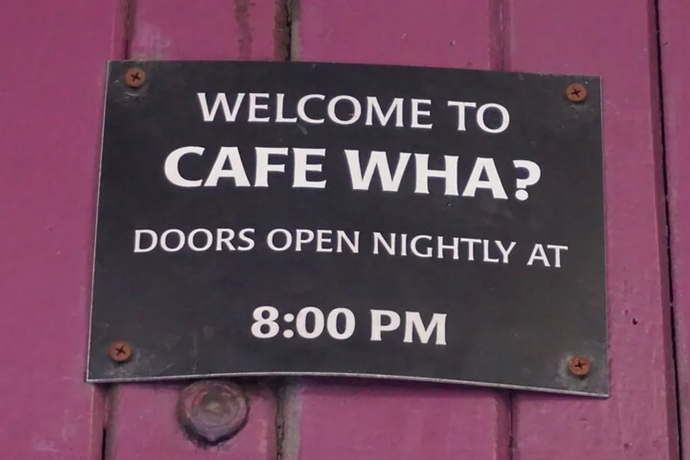 The image shows a black sign with white text that reads WELCOME TO CAFE WHA DOORS OPEN NIGHTLY AT 800 PM affixed to a purple wooden surface