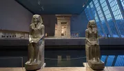 Two ancient Egyptian statues flank a water-filled channel leading to the Temple of Dendur, set against a sloping glass wall and a modern museum interior.
