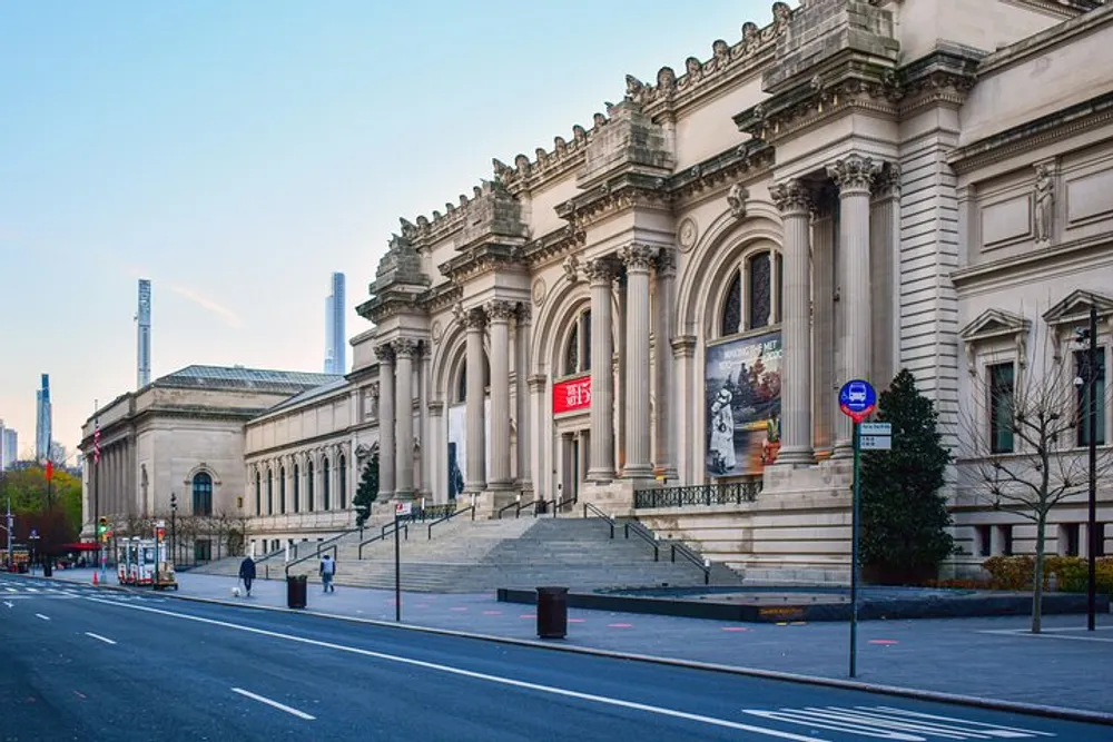 The image showcases the grand and ornate facade of the Metropolitan Museum of Art in New York City on a clear day with the street in the foreground exhibiting minimal traffic and activity