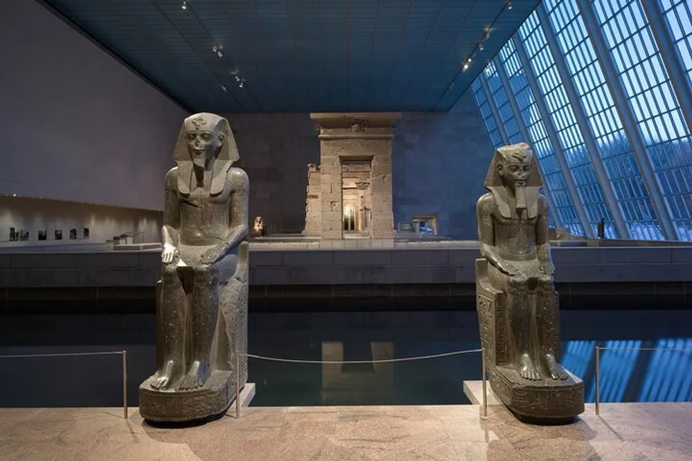 Two ancient Egyptian statues are displayed before a temple exhibit within a modern museum setting with a glass ceiling and minimalistic design