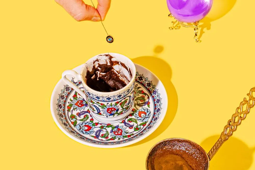 A hand is dropping a chocolate ball into a mug of hot chocolate on a patterned saucer set against a yellow background with a spoon and dangling purple balloons