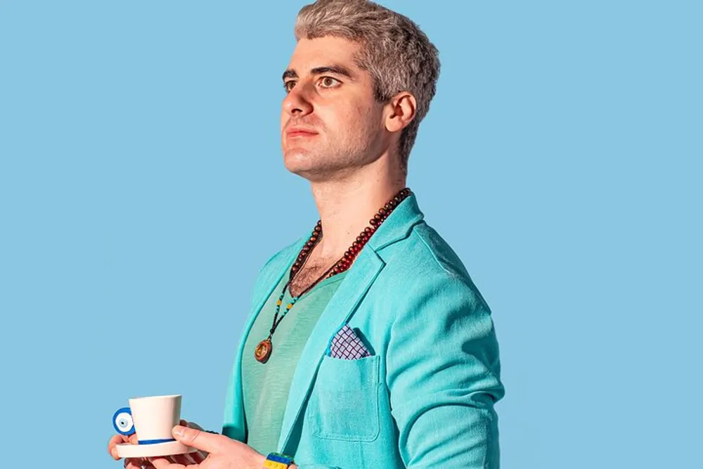 A person with gray hair wearing a teal blazer with a patterned pocket square multiple necklaces and holding a small cup and saucer stands in front of a blue background