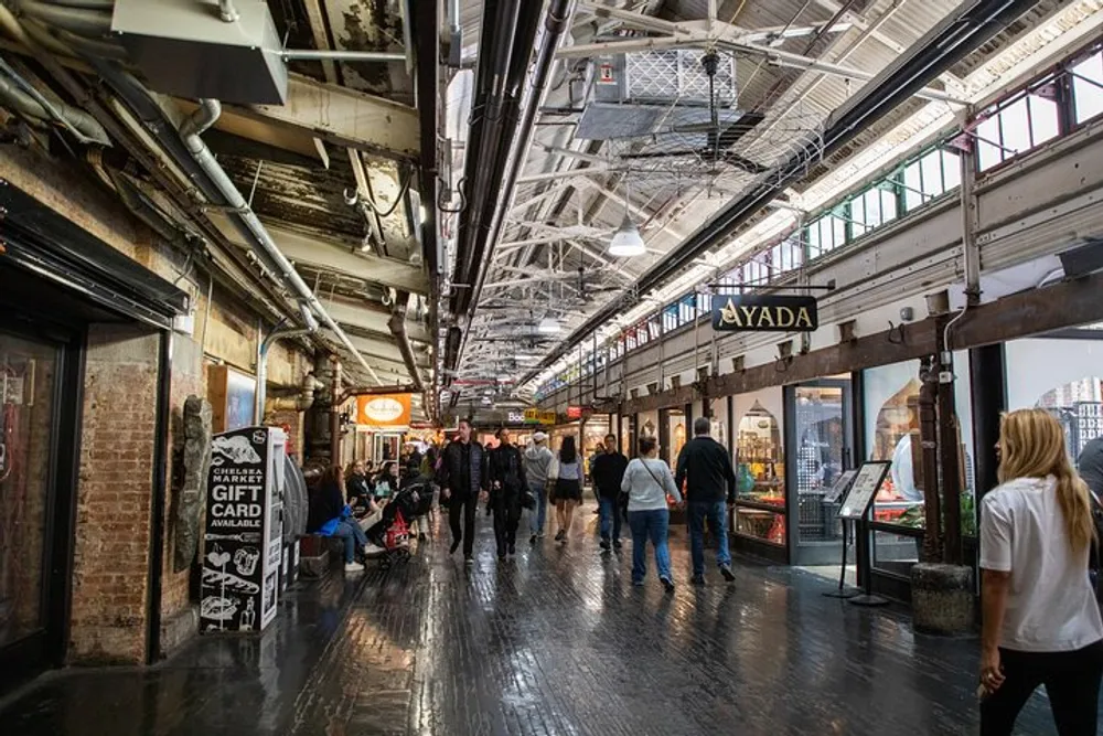 People are walking through a bustling indoor market with shops on either side and an industrial-style ceiling overhead