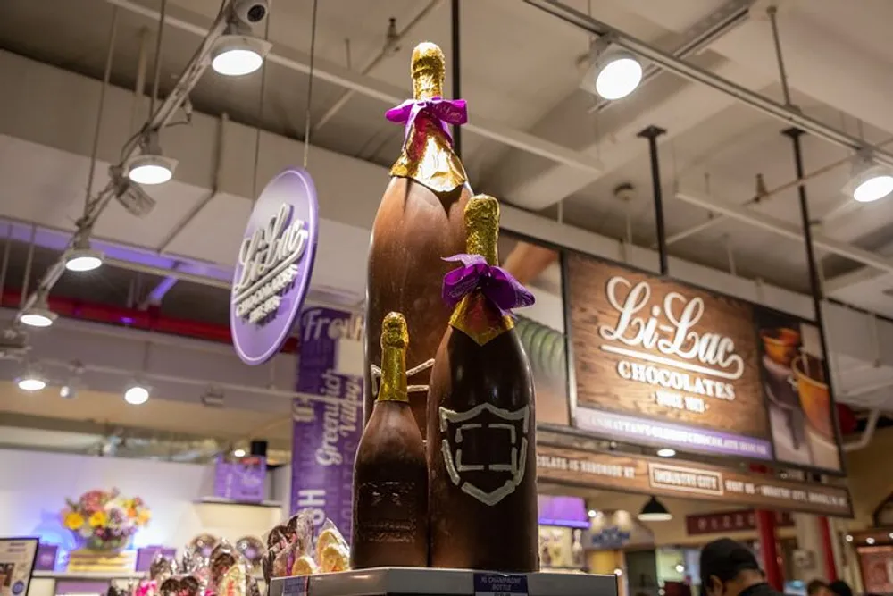 The image shows a large chocolate sculpture of a champagne bottle with a purple ribbon displayed in a store with other chocolate products and signage in the background