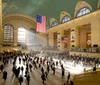 The image depicts a bustling Grand Central Terminal in New York City illuminated by natural light streaming through its large windows with the American flag prominently displayed above the crowded concourse