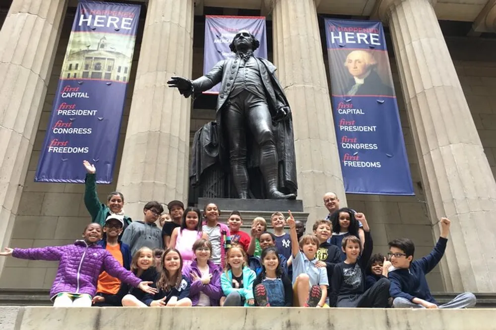 A group of children and adults pose happily on the steps in front of a large statue of a historical figure with educational banners hanging beside them
