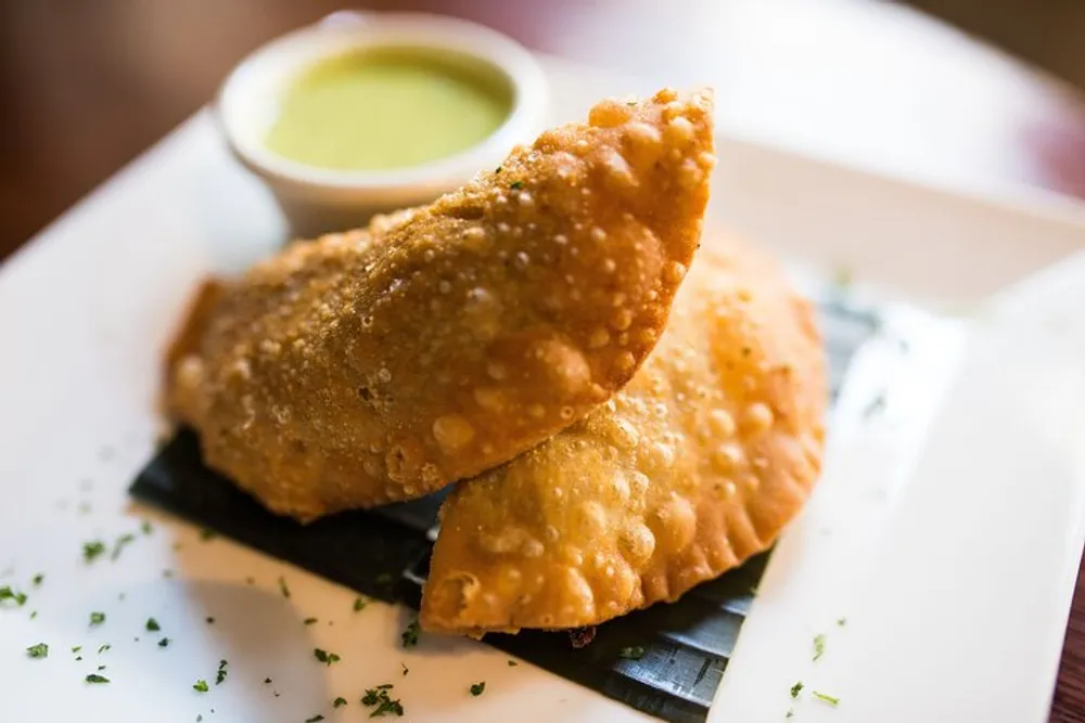 The image shows two golden-brown fried samosas on a white plate accompanied by a bowl of green chutney
