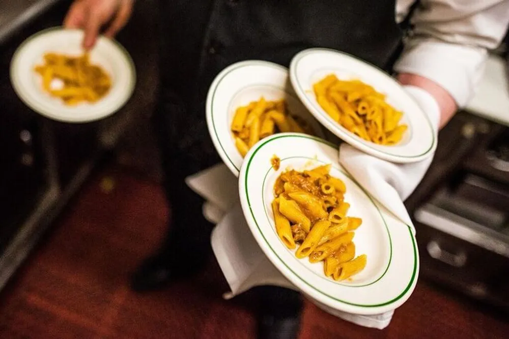 A server in a black uniform is carrying three plates of pasta possibly penne with a sauce stacked upon their arm